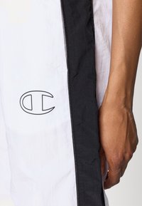 Krátký rukáv, bílý větruodolný kabát s černým bočním panelem a kontrastním logem. Tkanina má hladkou, mírně pomačkanou strukturu.