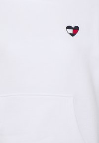 White sweatshirt with a small embroidered heart logo in navy, white, and red on the chest above a front pouch pocket.