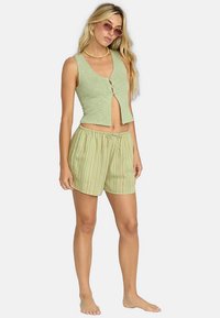 Žena oblečená v zelenom bezrukávovom crop topi s gombíkmi, pruhovaných šortkách so šnúrkou, slnečných okuliaroch a náhrdelníku, stojaca naboso na bielom pozadí.