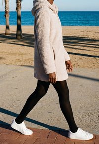 Personne portant un manteau duveteux beige clair, des collants noirs et des baskets blanches, marchant sur un chemin pavé près d'une plage de sable avec des palmiers.