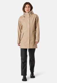 Veste étanche beige avec capuche, fermeture à boutons-pression, deux poches avant et pantalon noir. Conçue pour les activités de plein air et la protection.