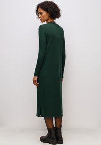 Abito lungo a maniche lunghe di colore verde scuro realizzato in un morbido materiale con spacchi laterali. Indossato con stivali neri alla caviglia. Design semplice e aderente.