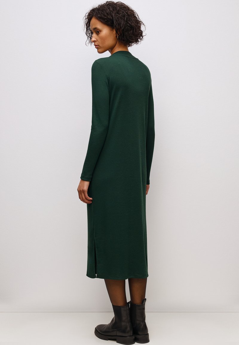 Abito lungo a maniche lunghe di colore verde scuro realizzato in un morbido materiale con spacchi laterali. Indossato con stivali neri alla caviglia. Design semplice e aderente.