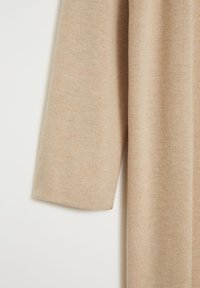 Tissu de pull beige à manches longues tricoté avec une texture douce visible, suspendu sur un fond blanc uni.