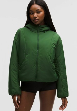 Naine seisab ning kannab rohelist kapuutsiga puffer-jakki esitaskutega ja musti lühikesi pükse tavalise tausta ees.