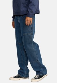 Pantalons cargo en denim bleu avec une coupe ample, featuring plusieurs poches, des coutures visibles et une texture subtile. Associé à des baskets noires.