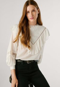 Blusa bianca con colletto e maniche a volant, caratterizzata da ricami floreali e dettagli in pizzo. Abbinata a jeans neri a vita alta e una cintura.