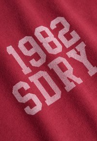Tessuto rosso con testo bianco in grassetto "1982 SORY" stampato sopra, mostrando la texture e lievi pieghe nel materiale.