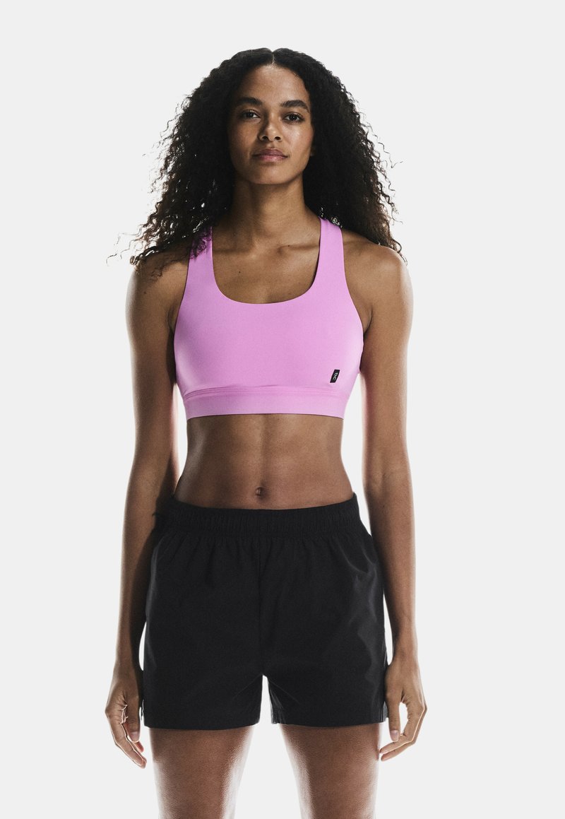 Sostén deportivo rosa con tirantes anchos, textura suave y escote cuadrado, combinado con pantalones cortos negros de cintura elástica.
