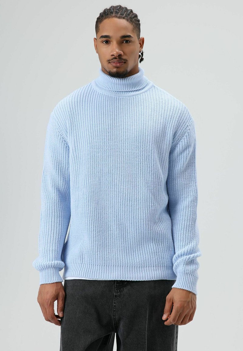 Pull à col roulé tricoté bleu clair avec texture côtelée et manches longues, associé à un jean foncé. Coupe ajustée avec poignets retroussés.
