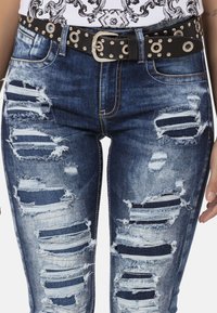 Versleten blauwe denim jeans met grote scheuren en rafelige randen. Gecombineerd met een zwarte riem met zilveren oogjes en een rechthoekige gesp.