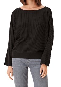 Femme portant un pull noir côtelé à manches longues et larges, associé à un jean gris clair coupé à la taille.