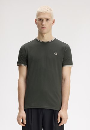 Jeune homme aux cheveux blonds courts portant un t-shirt ajusté vert foncé avec un col et des bords de manches rayés, debout devant un fond uni.