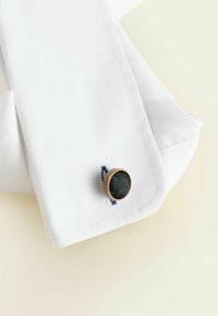 Vit skjorta med texturerad krage, knapphål i marinblå tråd och rund guldfärgad manschettknapp med svart tyg i mitten.