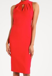 Robe rouge sans manches avec un col montant et une découpe en forme de goutte. Silhouette ajustée avec une texture lisse et un ourlet à hauteur du genou.