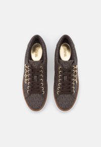 Un paio di sneakers marroni Michael Kors con motivo monogram, occhielli dorati, lacci marroni e charm "KORS" dorati alle estremità dei lacci.