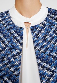 Tkany niebiesko-biały materiał z wzorem, z wysokim białym kołnierzem i małym otworem na szyję. Uzupełniony prostym srebrnym naszyjnikiem.