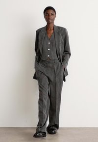 Costume gris à rayures avec un blazer ajusté et un pantalon à jambes larges. Tissu texturé avec des accents rouges subtils sur les rayures. Le mannequin se tient avec confiance.