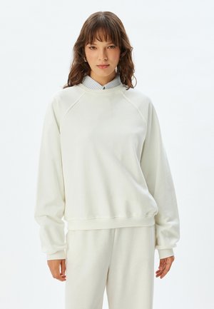 Valkoinen fleece-huppari, jossa on pyöreä kaula-aukko ja raglan-hihat. Hupparissa on ribattu hihansuu ja helma, ja se on kerroksittain päällepuettu kaulukselliselle, raidalliselle paidalle.