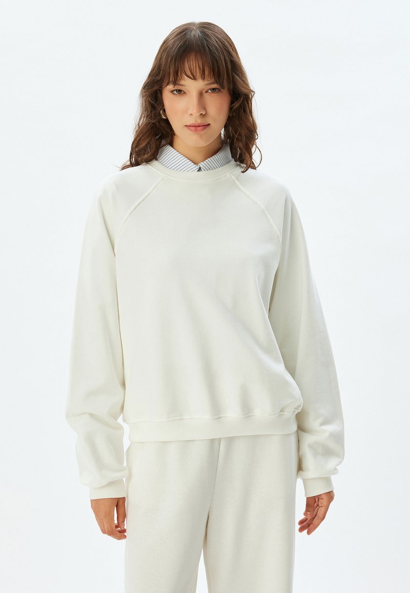 Sudadera de felpa blanca con cuello redondo y mangas raglán, que presenta puños y dobladillo acanalados, superpuesta a una camisa de rayas con cuello.