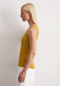 Senfgelbes, geripptes Tanktop mit breiten Trägern und figurbetontem Design. Kombiniert mit einem weißen Rock, seitlich dargestellt.