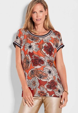 Blusenshirt mit Blumenmuster in Orange, mit schwarzen Akzenten und einem runden Ausschnitt. Kurze Ärmel und fließender Stoff fallen über metallisch goldene Hosen.