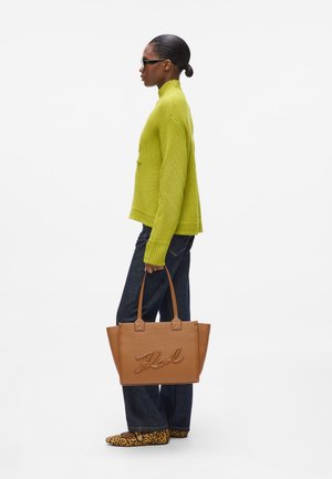Femme avec un chignon soigné, portant un pull vert, un jean foncé, des ballerines léopard, des lunettes de soleil et tenant un sac tote en cuir tan avec le logo "Karl".