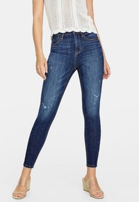 Jeans ajustados en denim de lavado oscuro con una textura desgastada y acentos rasgados. Combinados con una blusa de punto blanca y sandalias de cuña en color marrón.