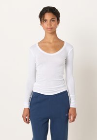 Valge pikakäise ja sügava V-kaelusega T-särk, kerge puuvillane kangas, sile tekstuur, siniste pükstega, millel on logoga detailid.