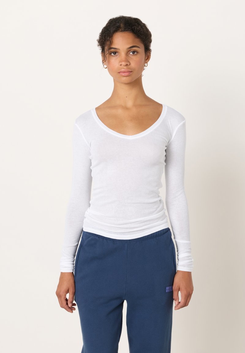 Valge pikakäise ja sügava V-kaelusega T-särk, kerge puuvillane kangas, sile tekstuur, siniste pükstega, millel on logoga detailid.