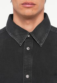 Chemise en denim noir avec un col pointu et des boutons-pression argentés, présentant une texture lisse et un léger dégradé sur le tissu.