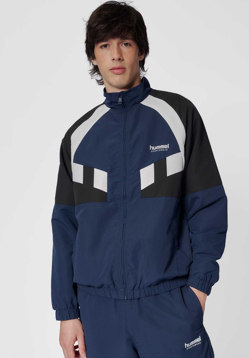 Młody mężczyzna w niebieskiej, czarnej i białej sportowej kurtce i pasujących spodniach Hummel, stojący z jedną ręką w kieszeni na jednolitym tle.