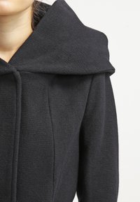 Black coat with a large hood, made of textured fabric. Features a fitted design with vertical seams and a single button closure.