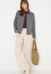 Koszula w czarno-białą kratę na purpurowym topie, beżowe spodnie o szerokich nogawkach oraz beżowe sneakersy z niebieskimi akcentami. Trzyma torebkę w kolorze khaki.