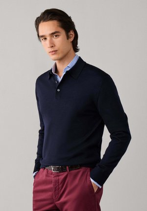 Joven con un polo azul marino de manga larga sobre una camisa de rayas azules y pantalones burdeos, con las manos en los bolsillos, de pie frente a un fondo liso.
