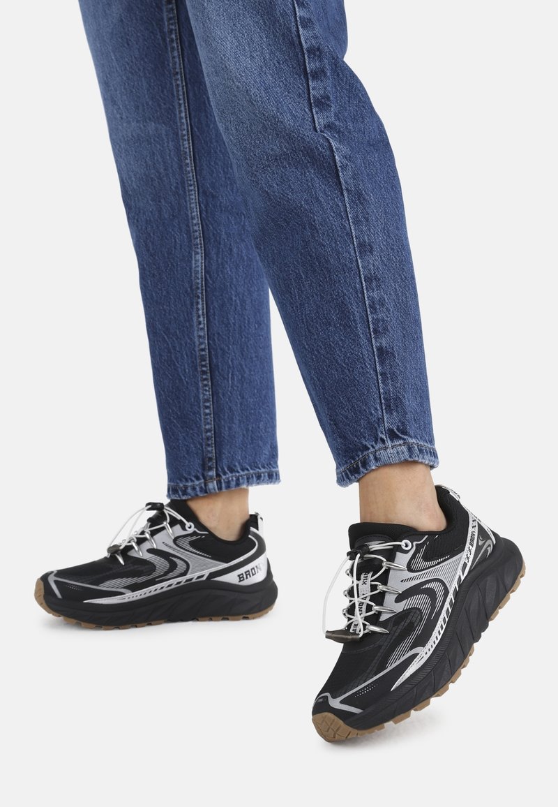 Baskets noires et grises avec un design texturé, dotées d'une semelle épaisse et de lacets élastiques, associées à un jean en denim bleu.