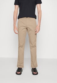 Beige Baumwollhosen im geraden Schnitt, mit Vordertaschen und Knopfverschluss. Kombiniert mit einem schwarzen T-Shirt und schwarzen Sandalen.