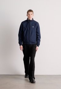 Στενή μπλε ελαφριά ζακέτα με ψηλό γιακά, κλείσιμο με φερμουάρ και πλαϊνές τσέπες, φορεμένη με μαύρο παντελόνι και παπούτσια, με καθαρό, μίνιμαλ σχεδιασμό.