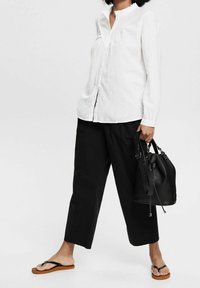 Camicia di lino bianca con colletto arruffato, abbinata a pantaloni larghi neri e infradito nere. Borsa nera con una texture morbida.