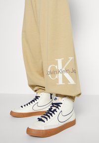 Crème hoge sneakers met marineblauwe accenten, gecombineerd met beige sweatpants met een wit "CK" logo en elastische boorden bij de enkels.