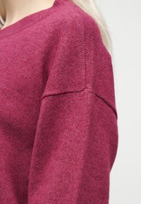 Γυναίκα που φοράει μαλακό, υφασμάτινο πουλόβερ με μακριά μανίκια σε βαθύ ματζέντα χρώμα με ορατές ραφές στον ώμο και στο πάνω μέρος του βραχίονα.