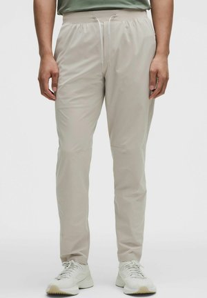 Mand iført lyse beige, slim-fit bukser med elastisk talje og snøre, kombineret med hvide sneakers og en grøn skjorte.