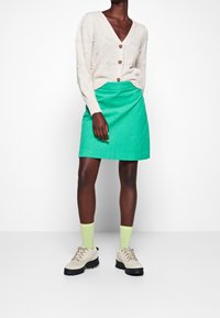 Cardigan beige strutturato con bottoni, abbinato a una gonna verde brillante. Calzini verde chiaro e scarpe beige chunky completano l'outfit.