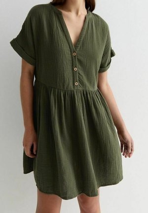 Femme portant une robe en tissu texturé vert olive à manches courtes, avec trois boutons en bois et une taille froncée, sur un fond uni clair.