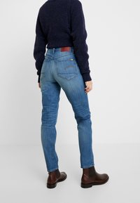 Blå denimjeans med en tajt passform, med bakfickor och subtila sömnadsdetaljer, tillsammans med bruna ankelkängor och en mörk tröja.