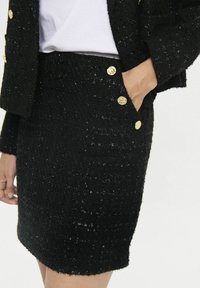 Schwarzer Tweed-Rock mit strukturierter Oberfläche, goldenen Knöpfen und seitlichen Taschen, taillierter Schnitt und Saum kurz über dem Knie.
