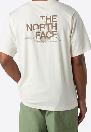 Personne portant un T-shirt blanc avec un graphique de montagne marron et le texte "THE NORTH FACE NEVER STOP EXPLORING" au dos, associé à un short vert.