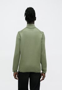 Zielony sweter dziany z kołnierzem, z prążkowanymi mankietami i pasem dolnym oraz ozdobnym przeszyciem na ramieniu. Noszony z ciemnymi dżinsami.