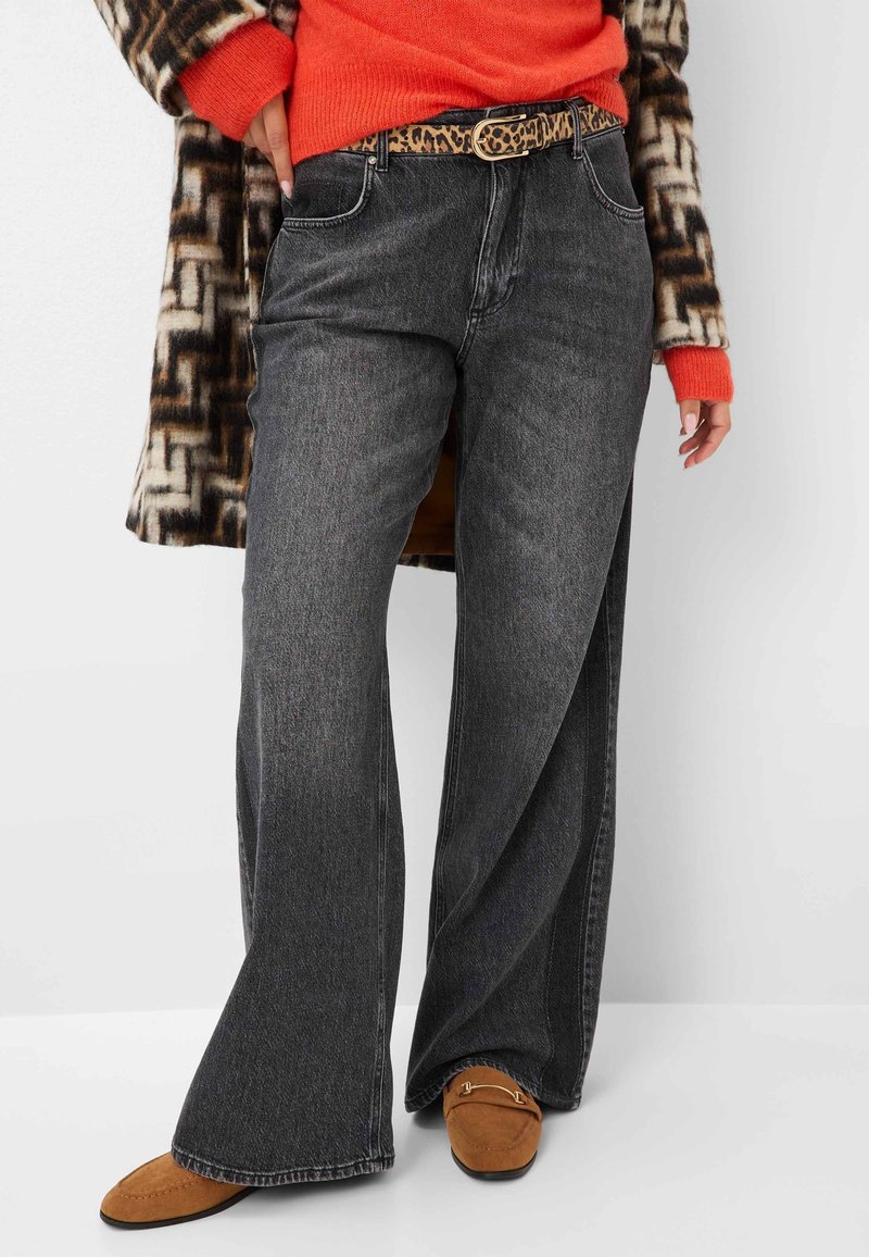 Personne portant un jean noir délavé à jambes larges, une ceinture imprimé léopard, un pull orange, des mocassins marron et un manteau à motif géométrique.