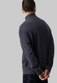 Dunkelgrauer Pullover mit hohem Kragen, strukturiertem Stoff, langen Ärmeln und lockerer Passform. Verfügt über Seitentaschen und gerippte Bündchen.